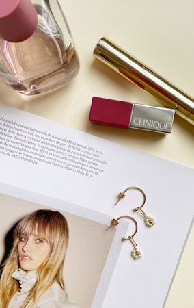 A magazine page is spread open featuring a fashion photograph of a model with long hair and a white outfit. Nearby, there are elegant gold earrings with pearl details placed on the page. Also visible are a pink-topped perfume bottle, a gold-colored cosmetic tube, and a pink Clinique lipstick on a light-colored surface.