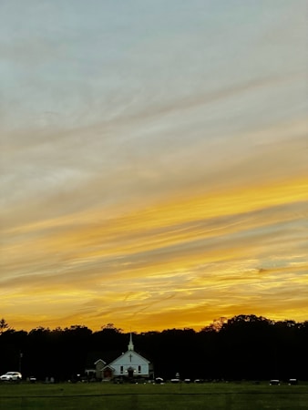 A picturesque sunset with vibrant yellow and orange hues fills the sky, casting a warm glow over a small white church with a steeple. The church is situated in a field with a background of dense, dark trees silhouetted against the colorful sky. A few cars are parked near the church, adding a sense of calm and stillness to the scene.