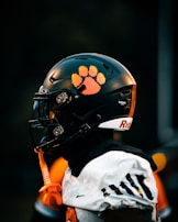 A football player is wearing a black helmet with a bright orange paw print decal on the side. The player is also wearing a white jersey with black and orange accents. An orange mouthguard attached to the helmet dangles down. The background is dark and out of focus, emphasizing the player's gear.