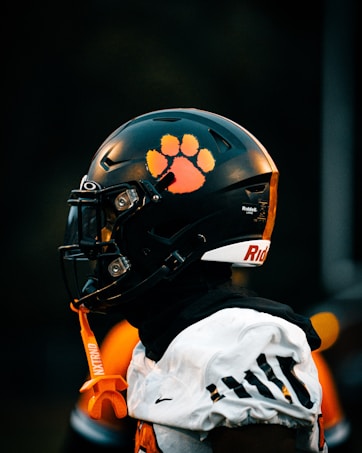 A football player is wearing a black helmet with a bright orange paw print decal on the side. The player is also wearing a white jersey with black and orange accents. An orange mouthguard attached to the helmet dangles down. The background is dark and out of focus, emphasizing the player's gear.