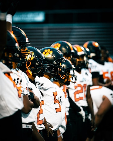 A line of American football players wearing black helmets with orange paw prints and white jerseys. The players are standing close together, their attention focused ahead, possibly at the start of a game or during a team huddle.