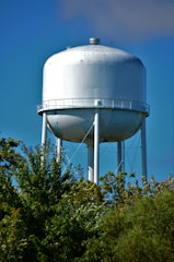 A tall, cylindrical water tower with a large tank is elevated above green trees. The tank is painted white and supported by several metal legs against a clear blue sky.