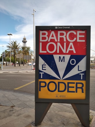 A street scene featuring an urban signboard with bold lettering and a geometrical design. The background includes a mix of palm trees, street signs, and a distinctive tower, suggesting a city landscape.