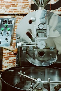 A large industrial coffee roaster with shiny metal surfaces, control knobs, and a gauge. The roaster appears to be in a rustic setting with a backdrop of red brick walls.