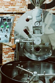 A large industrial coffee roaster with shiny metal surfaces, control knobs, and a gauge. The roaster appears to be in a rustic setting with a backdrop of red brick walls.