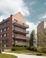 A modern residential building features multiple floors with balconies and a distinct brick façade. The structure is surrounded by lush greenery, including trees and well-manicured lawns. The sky is partly cloudy and adds a serene backdrop to the urban environment.
