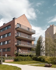 A modern residential building features multiple floors with balconies and a distinct brick façade. The structure is surrounded by lush greenery, including trees and well-manicured lawns. The sky is partly cloudy and adds a serene backdrop to the urban environment.
