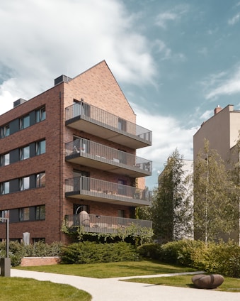 A modern residential building features multiple floors with balconies and a distinct brick façade. The structure is surrounded by lush greenery, including trees and well-manicured lawns. The sky is partly cloudy and adds a serene backdrop to the urban environment.