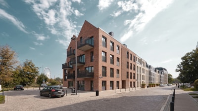A modern apartment building with a brick facade is situated at the corner of a cobblestone street. The building features balconies with black railings and large windows. In the background, a Ferris wheel is visible, along with trees lining the street. Several parked cars are seen along the road, and a few pedestrians are walking.