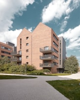 A modern apartment building with brick facades and multiple balconies. The building features a contemporary architectural design with angular roofs and large windows. Surrounding the structure are well-maintained shrubs and a paved walkway. The sky is partly cloudy, creating a calm and open atmosphere.