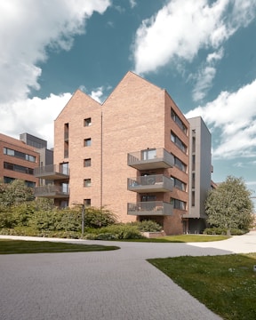 A modern apartment building with brick facades and multiple balconies. The building features a contemporary architectural design with angular roofs and large windows. Surrounding the structure are well-maintained shrubs and a paved walkway. The sky is partly cloudy, creating a calm and open atmosphere.