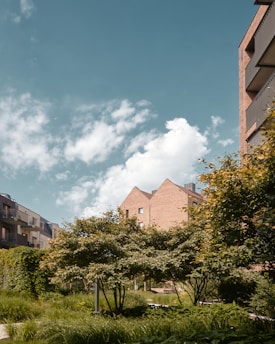 A modern residential building with balconies on the right is surrounded by lush, green trees and foliage. The brick structure of the building contrasts with the greenery, and the sky above is a vibrant blue with scattered white clouds.