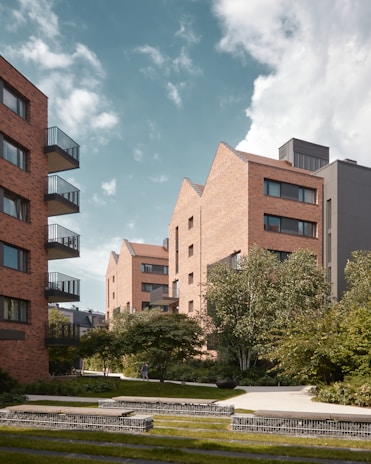 Several modern apartment buildings with brick facades are surrounded by lush greenery and landscaped gardens. The buildings have balconies and large windows. A paved walkway meanders through the well-maintained grass and trees. The sky is partly cloudy, providing a serene urban environment.