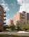 Several modern apartment buildings with brick facades are surrounded by lush greenery and landscaped gardens. The buildings have balconies and large windows. A paved walkway meanders through the well-maintained grass and trees. The sky is partly cloudy, providing a serene urban environment.