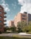 Several modern apartment buildings with brick facades are surrounded by lush greenery and landscaped gardens. The buildings have balconies and large windows. A paved walkway meanders through the well-maintained grass and trees. The sky is partly cloudy, providing a serene urban environment.