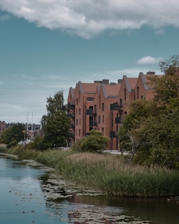 A row of modern brick apartment buildings lined along the edge of a calm waterway. The buildings have angular roofs and several balconies. Lush green trees and grass surround the water, which is partially covered by water lilies. The sky is overcast, adding a slightly moody ambience to the scene.
