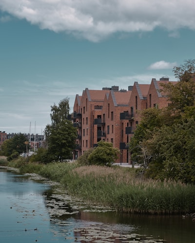 A row of modern brick apartment buildings lined along the edge of a calm waterway. The buildings have angular roofs and several balconies. Lush green trees and grass surround the water, which is partially covered by water lilies. The sky is overcast, adding a slightly moody ambience to the scene.