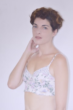A person with short, wavy hair is wearing a floral-patterned top. The background is plain and light in color, creating a soft, neutral setting. The individual holds a gentle, calm expression, with one hand touching their neck.