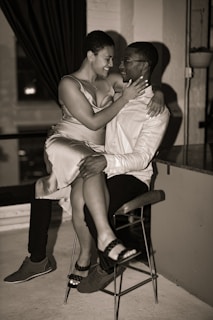 A couple is sitting together, closely interacting in an intimate and affectionate manner. The woman is seated on the man's lap, touching his face as they gaze at each other with smiles. The setting appears indoors with subdued lighting. The man wears a light shirt and dark pants, while the woman is dressed in a sleeveless dress.