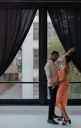 A couple is dancing near a large window with black curtains. The man is wearing a light-colored shirt and dark pants, while the woman is dressed in a bright orange dress and black high heels. They appear to be engaged in an intimate and joyful moment, looking into each other's eyes.