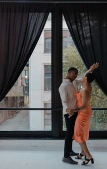 A couple is dancing near a large window with black curtains. The man is wearing a light-colored shirt and dark pants, while the woman is dressed in a bright orange dress and black high heels. They appear to be engaged in an intimate and joyful moment, looking into each other's eyes.