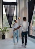 Two people are dancing together in a well-lit room with large windows. The atmosphere is casual and joyful, with one person laughing while they dance. There are dark curtains on the windows and a potted plant nearby, with sunlight streaming in through the windows.