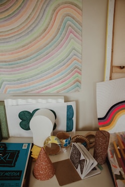 A desk area filled with various art projects and supplies, including a poster with curved, colorful lines, rolls of decorative tape, notebooks, and cardboard cones. There are sheets with abstract designs, with one featuring bold, wavy lines in shades of orange, yellow, and pink.