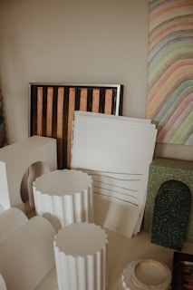 A variety of art supplies and decorative objects are neatly arranged. There are white sculptural elements resembling columns, a framed piece with vertical stripes, and sheets of paper with sketched lines. The background features a colorful, striped abstract painting.