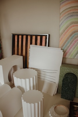 A variety of art supplies and decorative objects are neatly arranged. There are white sculptural elements resembling columns, a framed piece with vertical stripes, and sheets of paper with sketched lines. The background features a colorful, striped abstract painting.