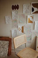A collection of abstract artworks is displayed on a wall, featuring a combination of organic shapes in black lines, colorful wavy patterns, and bold sections of gold and brown. A simple chair with a woven backrest and seat is positioned in front of the artwork. Additional framed art pieces are stacked against the wall.
