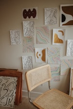 A collection of abstract artworks is displayed on a wall, featuring a combination of organic shapes in black lines, colorful wavy patterns, and bold sections of gold and brown. A simple chair with a woven backrest and seat is positioned in front of the artwork. Additional framed art pieces are stacked against the wall.