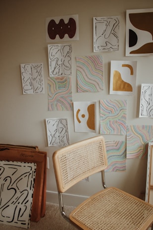 A collection of abstract artworks is displayed on a wall, featuring a combination of organic shapes in black lines, colorful wavy patterns, and bold sections of gold and brown. A simple chair with a woven backrest and seat is positioned in front of the artwork. Additional framed art pieces are stacked against the wall.
