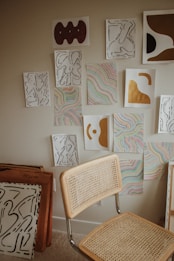 A collection of abstract artworks is displayed on a wall, featuring a combination of organic shapes in black lines, colorful wavy patterns, and bold sections of gold and brown. A simple chair with a woven backrest and seat is positioned in front of the artwork. Additional framed art pieces are stacked against the wall.