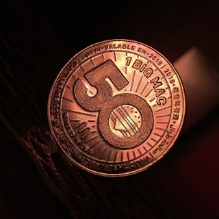 A close-up view of a coin-like token that has the number 50 prominently displayed in the center with a burger illustration. The words '1 BIG MAC' and various other texts in multiple languages can be seen around the edges. The token has a metallic sheen, and it is resting on a dark textured surface.