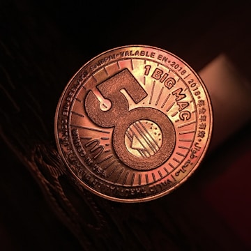 A close-up view of a coin-like token that has the number 50 prominently displayed in the center with a burger illustration. The words '1 BIG MAC' and various other texts in multiple languages can be seen around the edges. The token has a metallic sheen, and it is resting on a dark textured surface.
