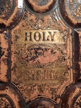 An ornate cover of a worn and aged book titled 'Holy Bible' with intricate golden designs and decorations. The leather cover shows significant signs of wear and tear, with sections of the material stripped away revealing a darker underlying surface. There are elaborate floral patterns surrounding the central title.