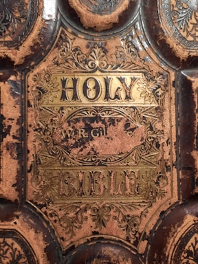 An ornate cover of a worn and aged book titled 'Holy Bible' with intricate golden designs and decorations. The leather cover shows significant signs of wear and tear, with sections of the material stripped away revealing a darker underlying surface. There are elaborate floral patterns surrounding the central title.