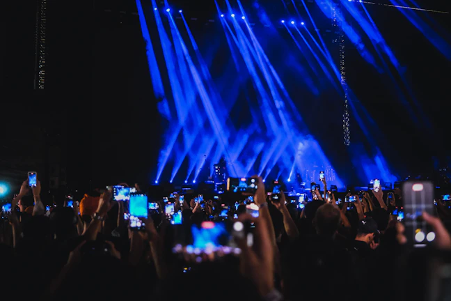 A large crowd of people holding up smartphones with their screens lit, capturing or recording an entertainment event on a stage illuminated by prominent blue spotlights.