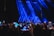 A large crowd of people holding up smartphones with their screens lit, capturing or recording an entertainment event on a stage illuminated by prominent blue spotlights.