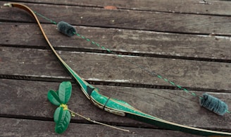 A green recurve bow is laid on a weathered wooden surface alongside some green leaves with small yellow flowers.