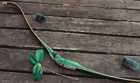 A green recurve bow is laid on a weathered wooden surface alongside some green leaves with small yellow flowers.
