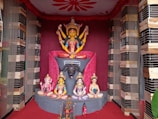 A colorful and intricate display of Hindu deities, featuring a central figure with multiple arms seated on a lion atop an ornate pedestal. Surrounding the main deity are smaller statues of other gods in various poses. The background is decorated with red drapery and wooden framing.