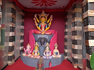 A colorful and intricate display of Hindu deities, featuring a central figure with multiple arms seated on a lion atop an ornate pedestal. Surrounding the main deity are smaller statues of other gods in various poses. The background is decorated with red drapery and wooden framing.