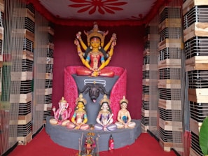 A colorful and intricate display of Hindu deities, featuring a central figure with multiple arms seated on a lion atop an ornate pedestal. Surrounding the main deity are smaller statues of other gods in various poses. The background is decorated with red drapery and wooden framing.