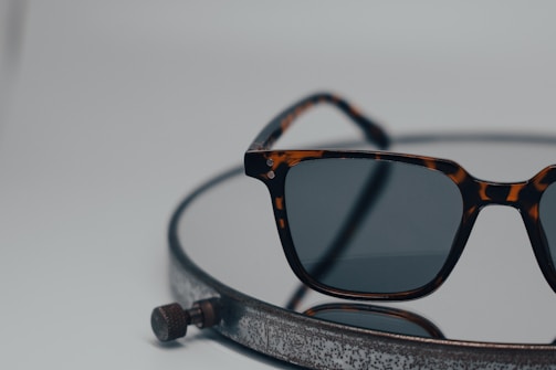 A pair of tortoiseshell sunglasses with dark lenses rests on a circular, metallic surface that has a rustic appearance. The background is out of focus, creating a minimalist and modern aesthetic.
