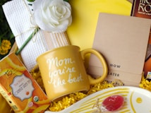 A yellow gift mug with the words 'Mom, you're the best' surrounded by a variety of items including a white rose, a box of orange green tea, a set of brown notepads labeled 'Now', 'Later', 'Eventually', and a plate with a red lollipop. The background is filled with yellow shredded paper, contributing to a celebratory feel.