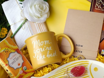 A yellow gift mug with the words 'Mom, you're the best' surrounded by a variety of items including a white rose, a box of orange green tea, a set of brown notepads labeled 'Now', 'Later', 'Eventually', and a plate with a red lollipop. The background is filled with yellow shredded paper, contributing to a celebratory feel.