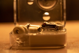 A pair of wireless earbuds rests elegantly in a transparent, compact charging case. The earbuds have a sleek, modern design with metallic and white accents. The lighting creates a warm, ambient glow, highlighting the reflective surfaces of the earbuds.