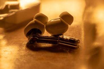 A pair of wireless earbuds rests on a reflective surface. The lighting is warm and highlights the smooth, glossy texture of the earbuds. Surrounding the earbuds, there is a blurred background that adds depth to the image.