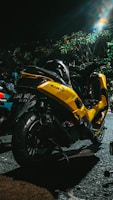 A yellow motorcycle is parked on a dimly lit street. The vehicle has a sleek design with the brand name 'NMax' visible on the side. It is positioned near some greenery, and there are other vehicles nearby. The scene is captured at night with artificial lighting creating a reflective effect on the bike's surface.